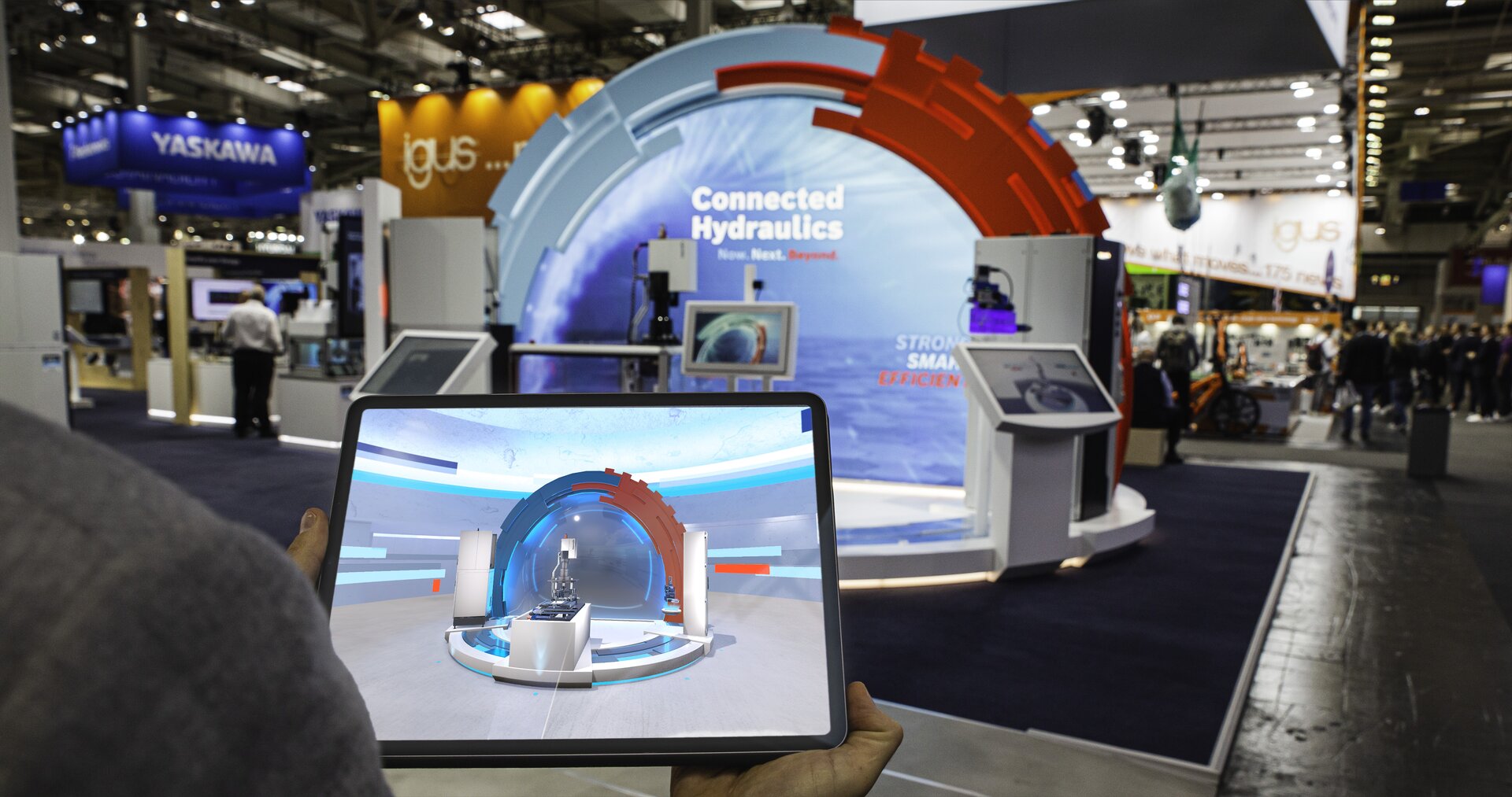 Blick auf eine Messehalle mit dem Rexroth-Stand „Connected Hydraulics“. Im Vordergrund hält eine Person ein Tablet mit einer VR-Visualisierung des realen Standes.