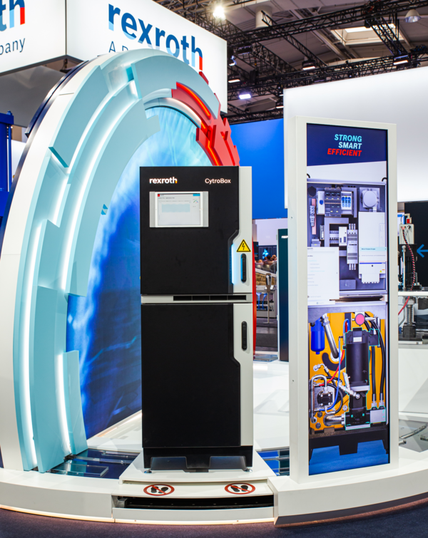 Messeexponat mit der „CytroBox“ von Rexroth vor einer inszenierten Rückwand. Rechts daneben ein Infodisplay mit technischen Abbildungen.