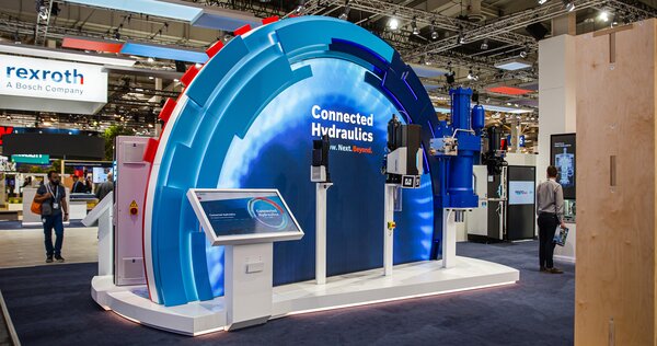 Messestand mit halbrunder Leuchtinstallation, die verschiedene Rexroth-Hydrauliksysteme präsentiert. Großes Schild: „Connected Hydraulics – Now. Next. Beyond.“