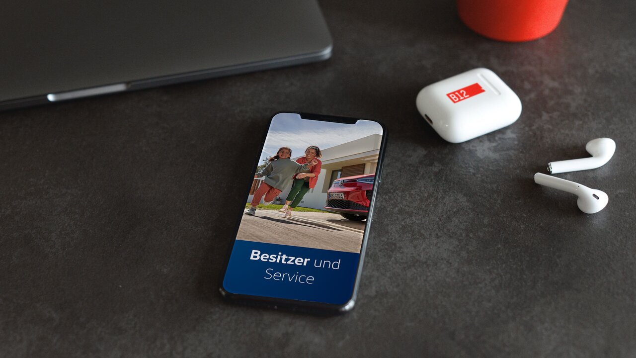 Smartphone auf Tisch zeigt VW-Service-App mit spielenden Kindern vor rotem Auto. Daneben AirPods mit „B12“-Label.