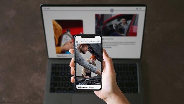 Hand hält Smartphone mit VW-Servicebild. Im Hintergrund ein Laptop mit passender Website geöffnet.
