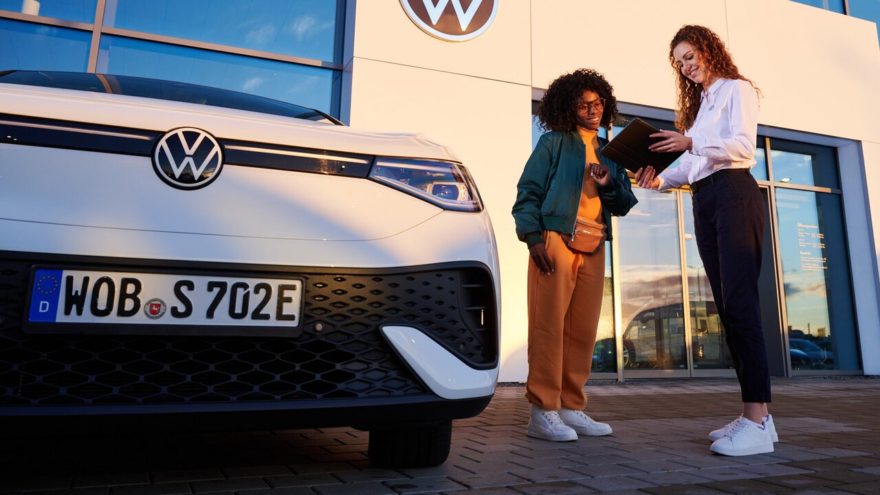 Zwei Frauen stehen vor einem weißen Volkswagen-Elektroauto und betrachten gemeinsam ein Tablet.
