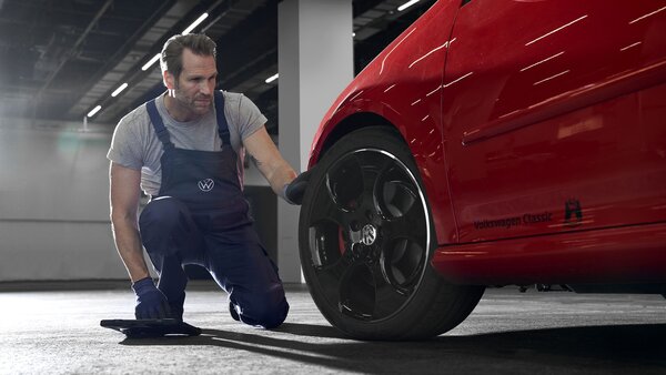 Mechaniker kniet vor rotem VW-Klassiker und kontrolliert das Vorderrad. Werkstattszene mit starker Ausleuchtung.