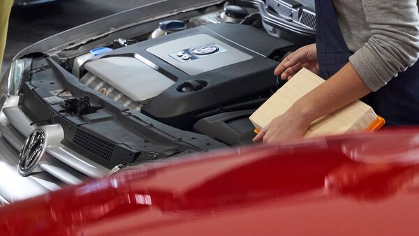 Nahaufnahme eines geöffneten Motorraums mit VW-Logo. Mechaniker:in hält Luftfiltereinsatz in der Hand.