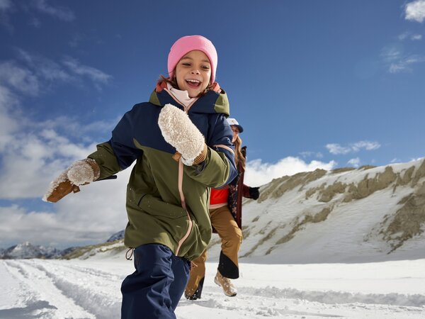 Kind in Winterkleidung rennt fröhlich im Schnee, Eltern folgen im Hintergrund. Blauer Himmel und verschneite Berglandschaft.